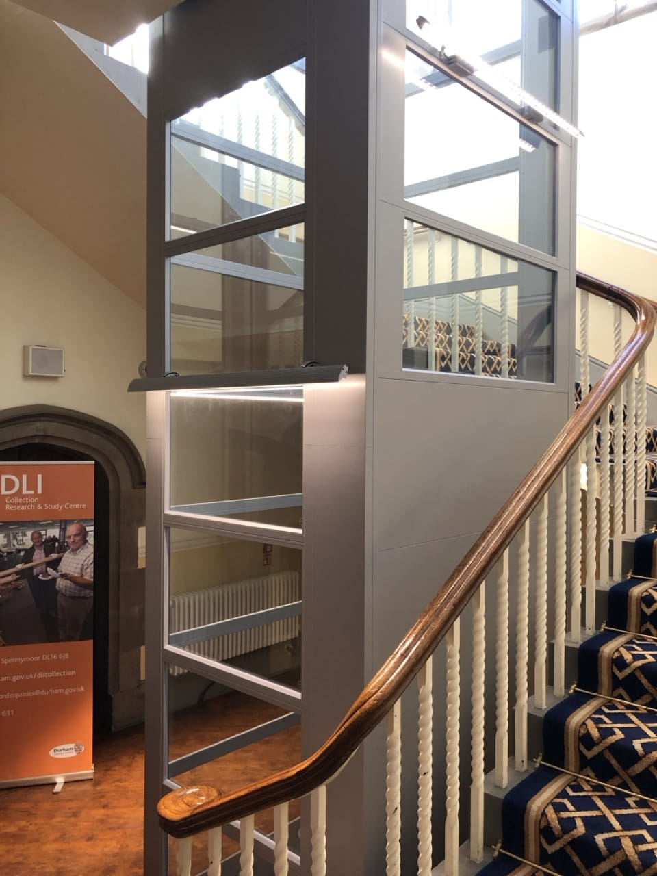 Modern grey and glass platform lift inside a building alongside a carpeted staircase with wooden railing. Nearby is a promotional poster on the floor.
