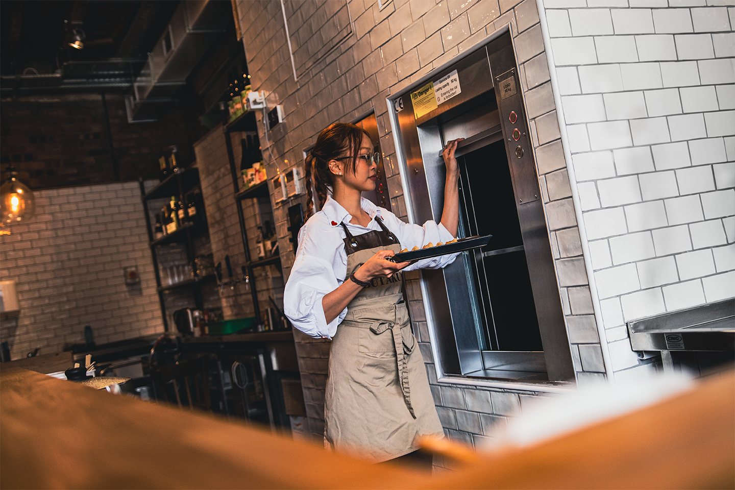 A woman in a white shirt and brown apron stands at an open dumbwaiter lift holding a tray of food. The kitchen has white tiles and industrial decor.