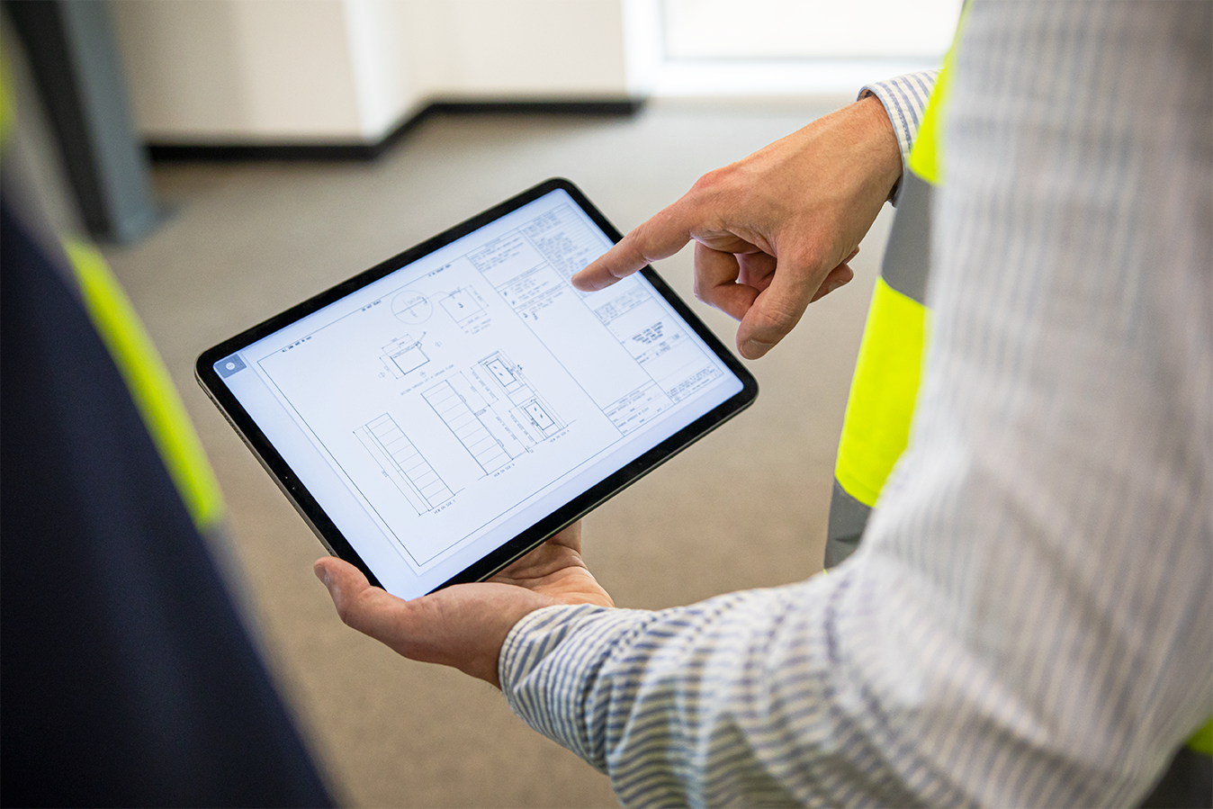 A person in a yellow safety vest points at architectural blueprints displayed on a tablet, suggesting a construction or design discussion in progress.